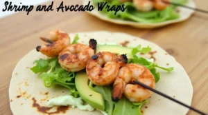 Shrimp and Avocado Wraps, Grilled shrimp skewers on a tortilla with fresh avocado slices, lettuce, and a drizzle of sauce, served on a wooden table.